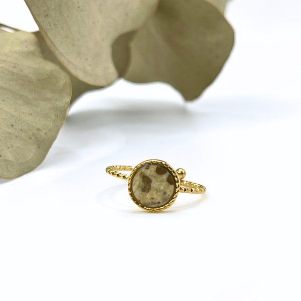 Bague délicate en acier inoxydable doré avec pierre beige, posée sur un fond végétal.