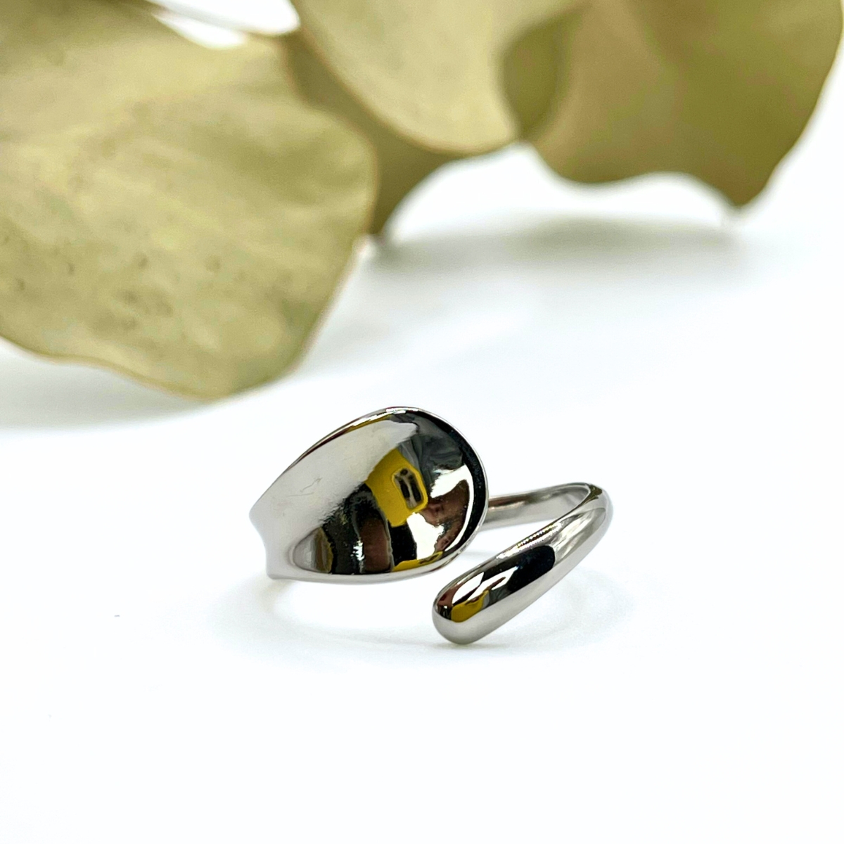 Bague en acier inoxydable argenté avec une forme organique et asymétrique, posée sur un fond végétal.