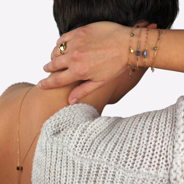 Une main ornée de délicats bracelets en acier inoxydable doré, avec des détails raffinés tels que des perles et des motifs étoilés. La personne porte également une bague dorée imposante et élégante, tout en touchant sa nuque, dévoilant un collier discret dans son dos. L’ensemble respire la sophistication et la douceur, parfaitement assorti à un pull tricoté beige.