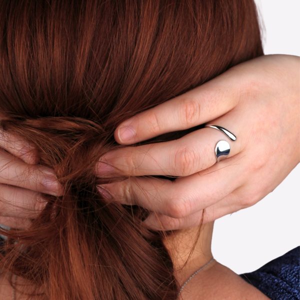 Une main aux doigts fins qui retient des cheveux roux, ornée d’une bague en acier inoxydable argentée au design asymétrique. La simplicité et le brillant de la bague captent l’attention, apportant une touche de modernité à ce geste naturel et spontané.