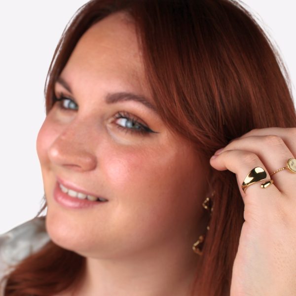 Un portrait en gros plan d’une femme aux cheveux roux, souriante, avec une bague dorée asymétrique à son doigt, accompagnée d’une autre bague fine et d’un bijou discret en arrière-plan. La lumière douce met en valeur la brillance du métal doré, soulignant l’allure chaleureuse et élégante du modèle.